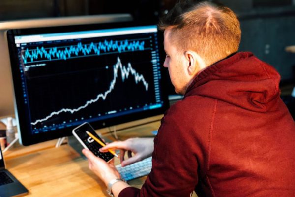 Stock Trading - man holding black smartphone with flat screen monitor in front