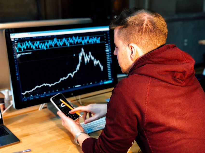 Stock Trading - man holding black smartphone with flat screen monitor in front