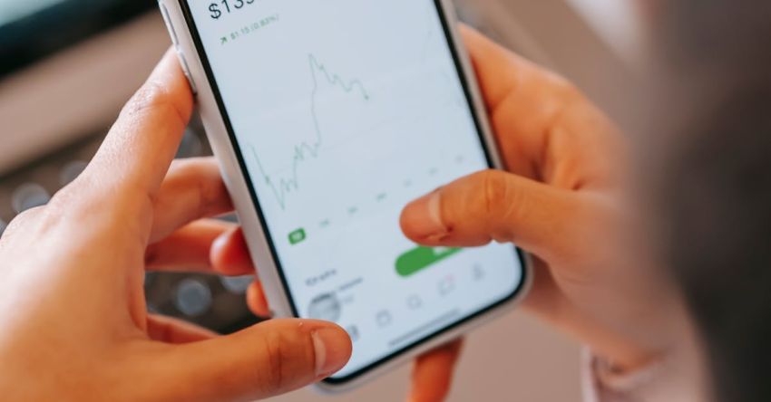 Stock Trading - High angle of crop anonymous female broker using app on cellphone with graphic on screen against netbook at home