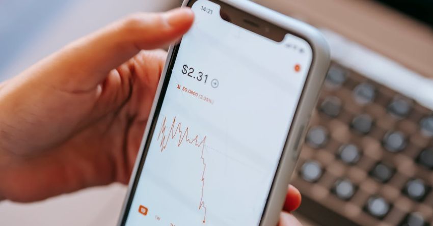 Stock Trading - High angle of crop anonymous dealer using financial exchange application on cellphone with chart on screen against laptop