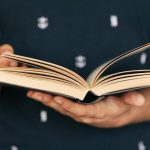 Literacy - Anonymous male in dark tee shirt reading interesting book while standing in front of light wall