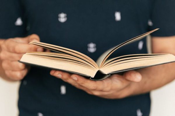 What Is the Importance of Financial Literacy Literacy - Anonymous male in dark tee shirt reading interesting book while standing in front of light wall