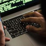 Fraud - Unrecognizable hacker with smartphone typing on laptop at desk