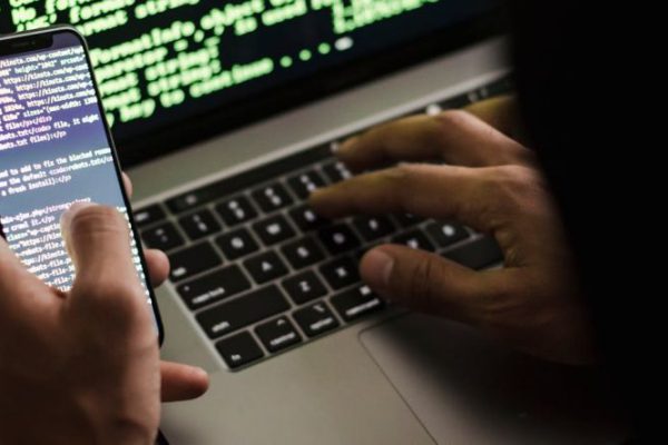 Fraud - Unrecognizable hacker with smartphone typing on laptop at desk